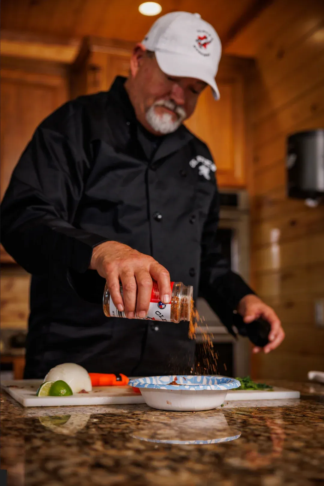 Cleve in a black chef coat and Cleve's ball cap shaking seasoning over fresh ingredients at the kitchen counter